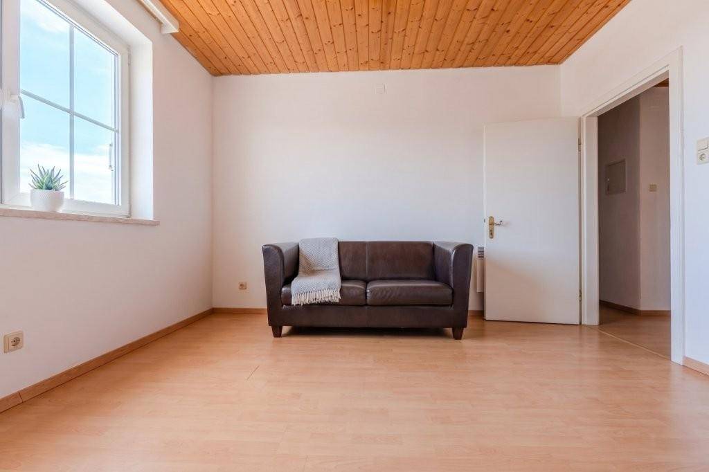 Gemütliches Wohnzimmer mit braunem Sofa, hellem Laminatboden und Holzdecke, durchflutet von Tageslicht.