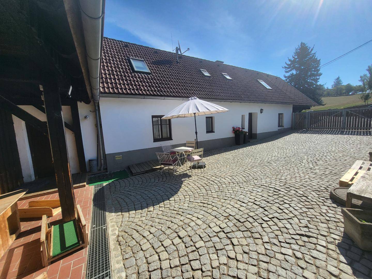 Ansicht des Hauses vom gepflasterten Hof aus, mit Dachfenstern und einem kleinen Sitzbereich unter einem Sonnenschirm.