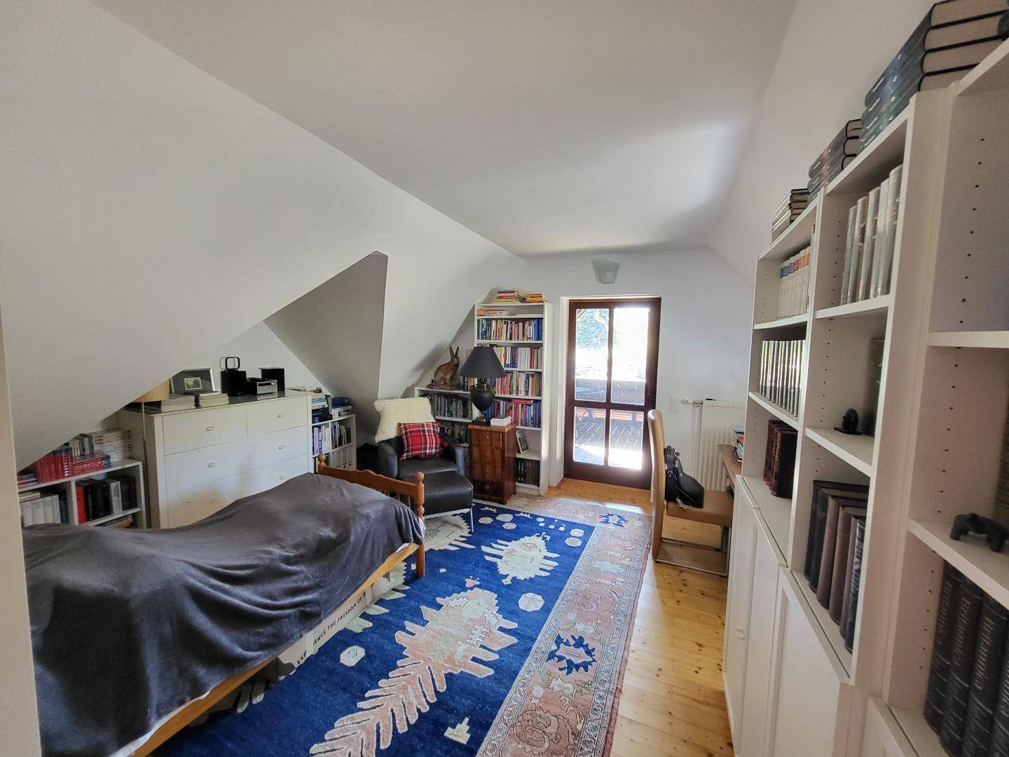 Helles Zimmer mit Bücherregalen, Einzelbett und Zugang zu einem Balkon, ideal als Schlaf- oder Arbeitszimmer.