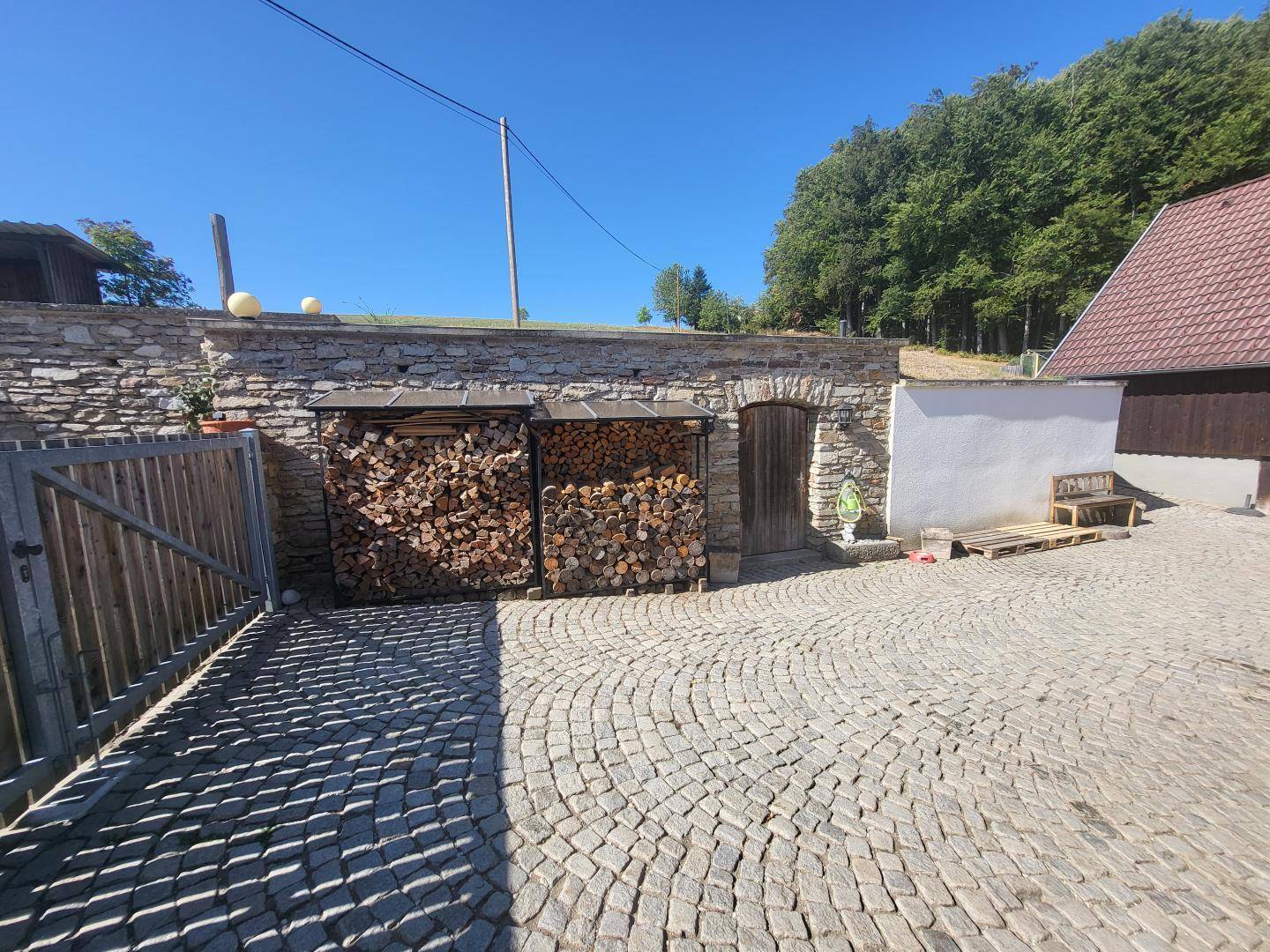 Gepflasterter Bereich neben einer Steinmauer mit gestapeltem Brennholz und einem rustikalen Holztor.