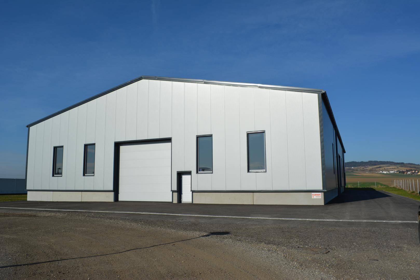 Moderne Gewerbehalle mit weißer Metallfassade, großem Rolltor und weitem Blick auf die Landschaft.