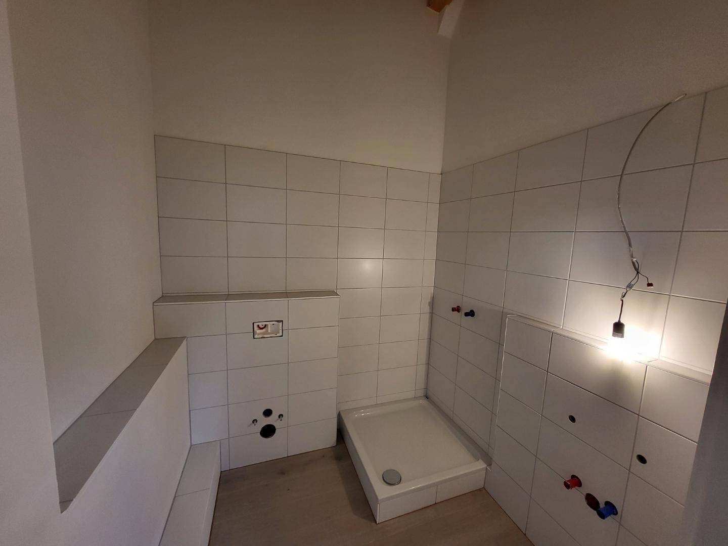 Unfertiges Badezimmer mit weißen Fliesen, Duschwanne und sichtbaren Sanitäranschlüssen.