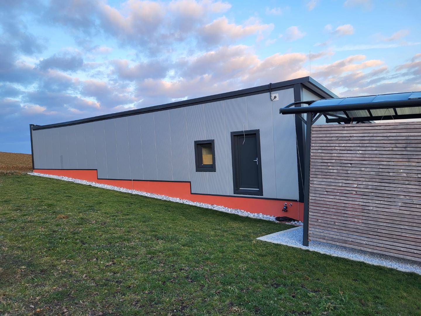 Modernes, rechteckiges Gebäude mit grauer Fassade und orangefarbenem Sockel in ländlicher Umgebung.
