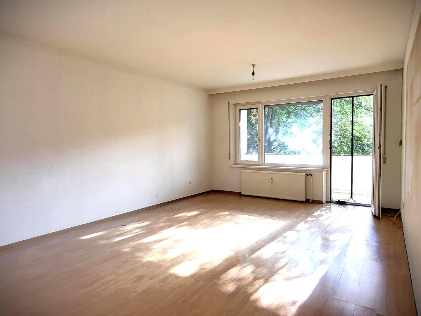 Helles Zimmer mit Holzfußboden, großem Fenster und Zugang zu einem Außenbereich.