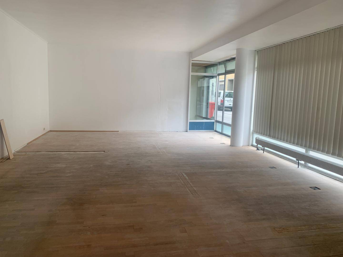 Großer, heller Raum mit Holzparkettboden, weißen Wänden und vertikalen Jalousien an den Fenstern.
