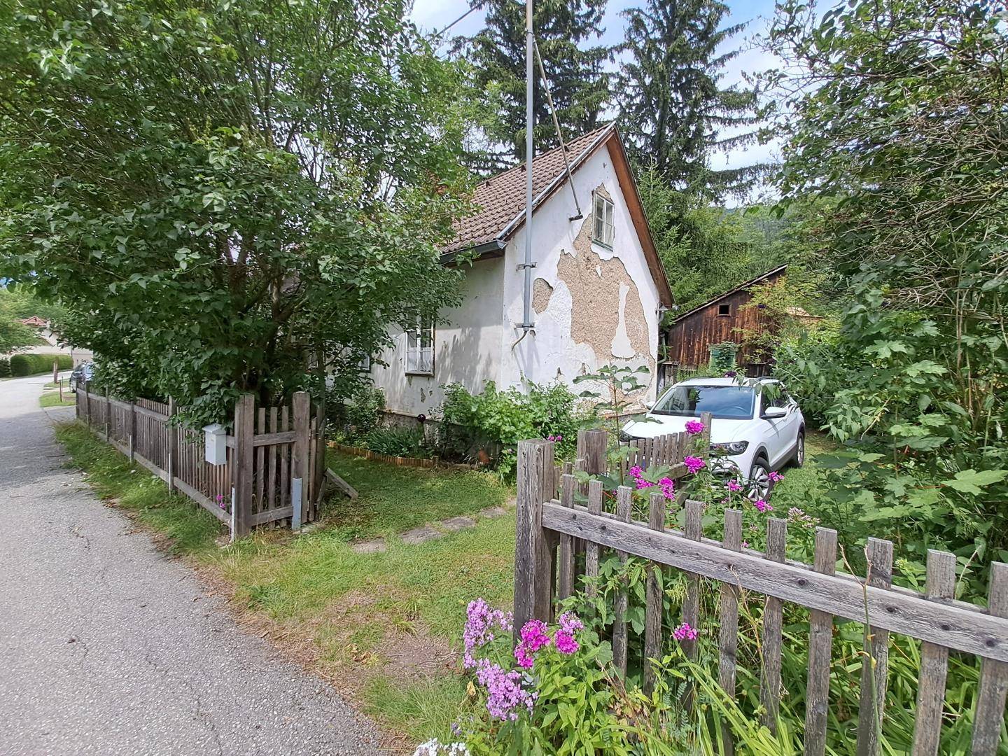 Ländliches Haus mit abblätternder Fassade, Holzzaun und parkendem Auto.