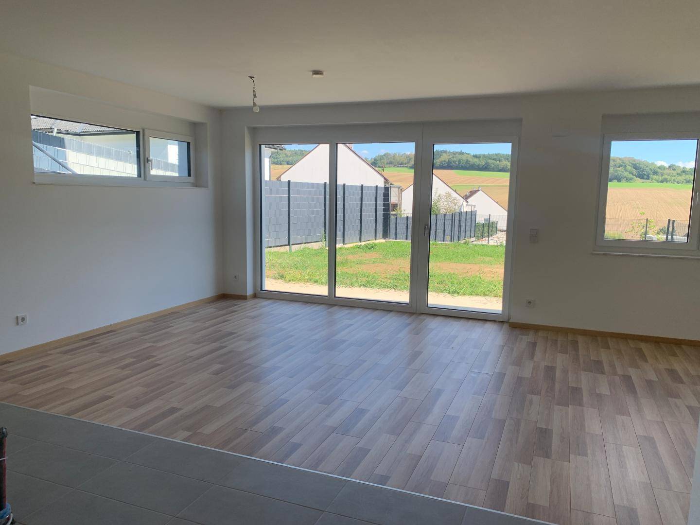 Lichtdurchfluteter Wohnbereich mit Laminatboden und Blick ins Grüne durch große Fenster.
