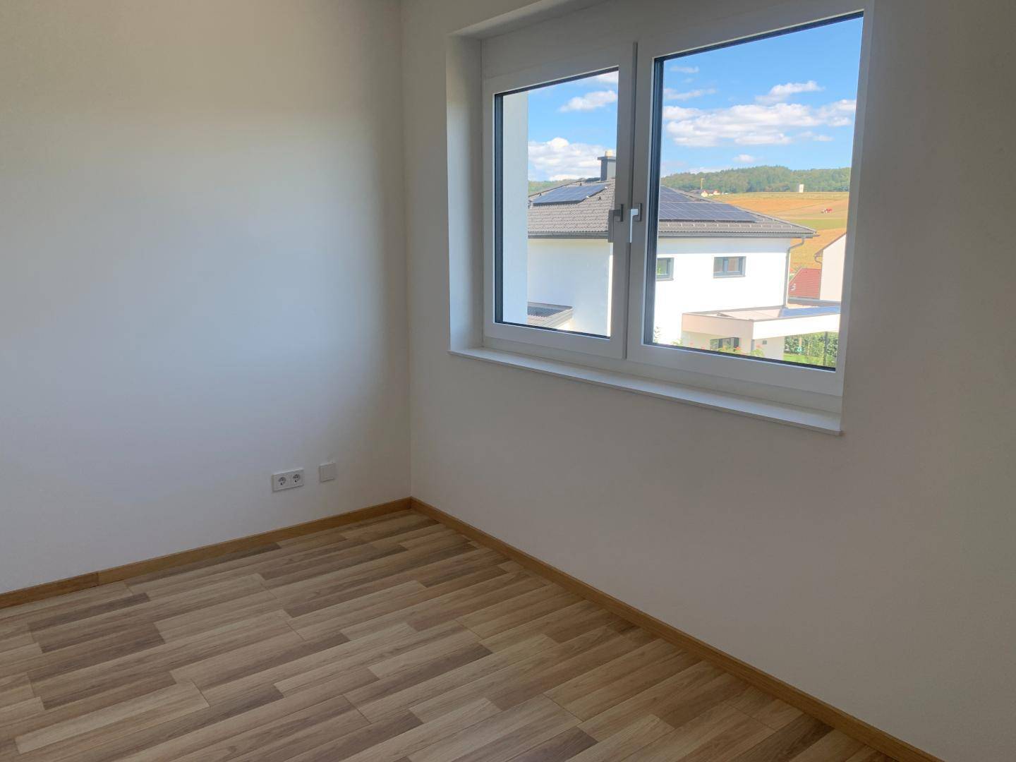 Leeres Zimmer mit Laminatboden und Fenster, das einen Blick auf benachbarte Häuser bietet.
