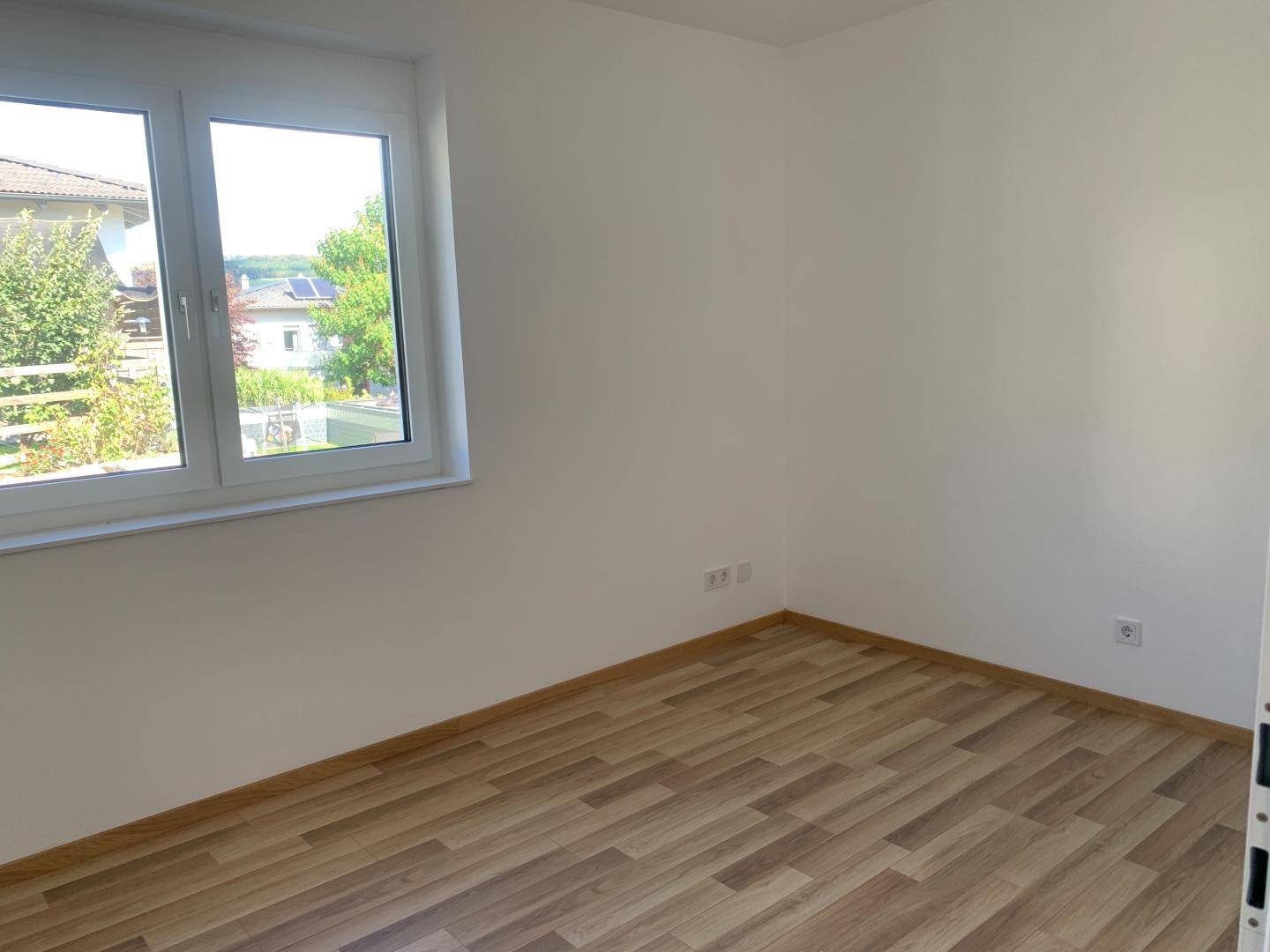 Leeres Zimmer mit Laminatboden und großem Fenster, das viel Tageslicht hereinlässt.