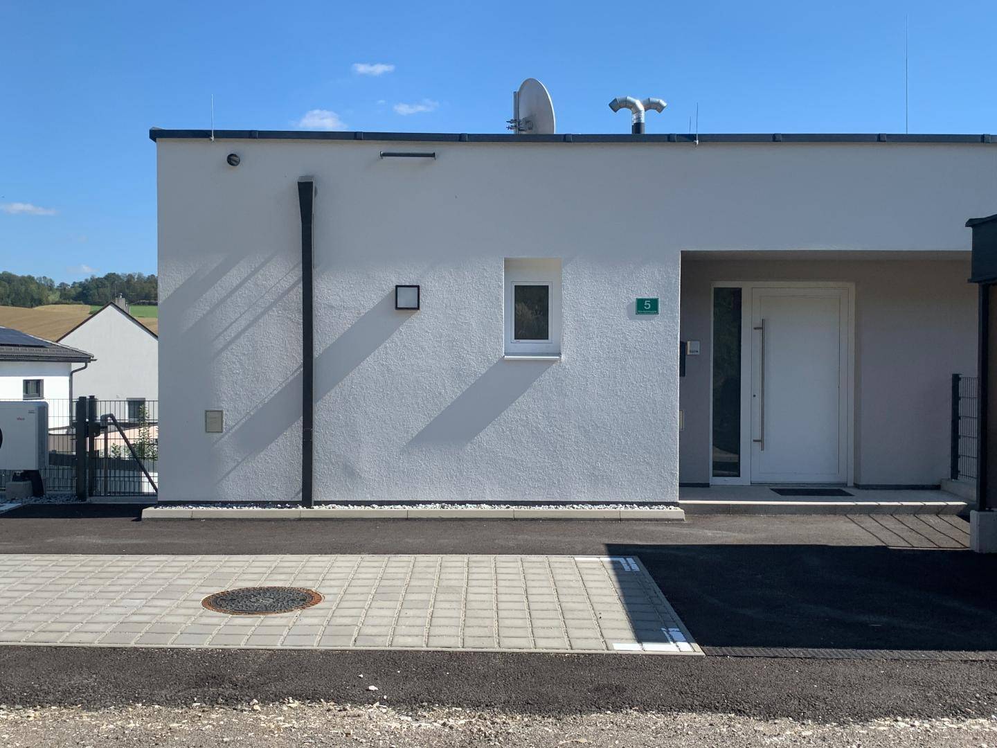 Moderner Hauseingang mit schlichter Fassade und gepflastertem Vorplatz unter blauem Himmel.