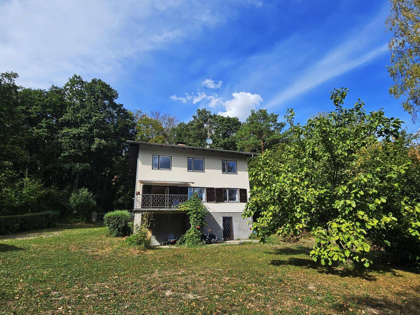 Freistehendes Haus mit Balkon und Rollläden, eingebettet in eine grüne Umgebung mit altem Baumbestand.