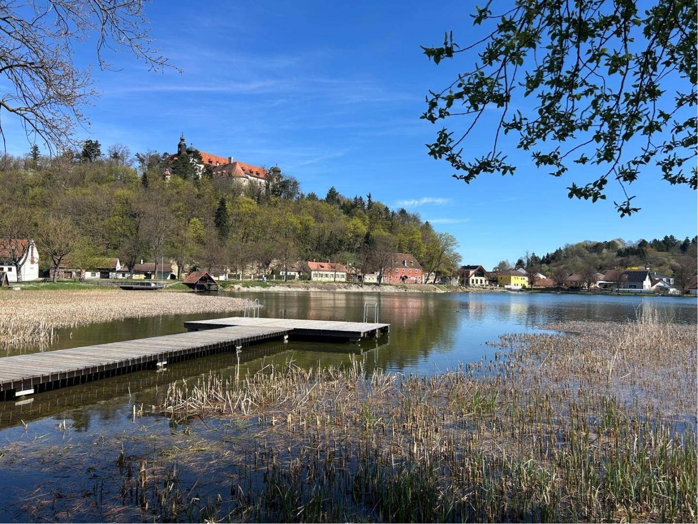 Malerischer See mit Steg und historischem Gebäude im Hintergrund unter blauem Himmel.