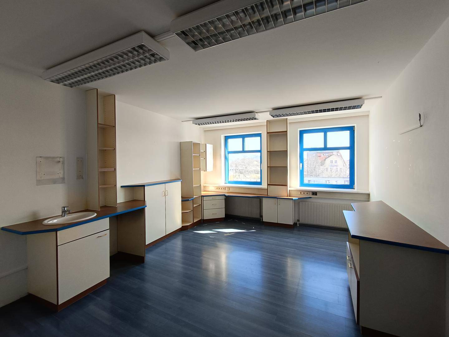 Büroraum mit Einbaumöbeln, Waschbecken und blauen Fensterrahmen, gut beleuchtet und funktional.