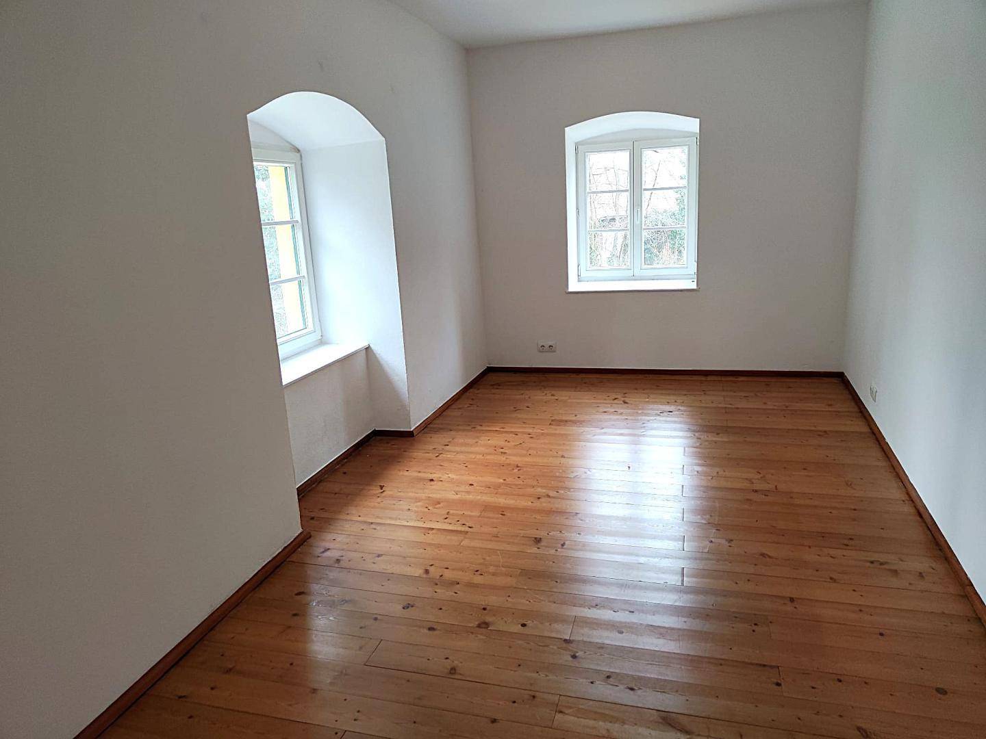 Leeres Zimmer mit Holzdielenboden und zwei Fenstern, die viel Tageslicht hereinlassen.