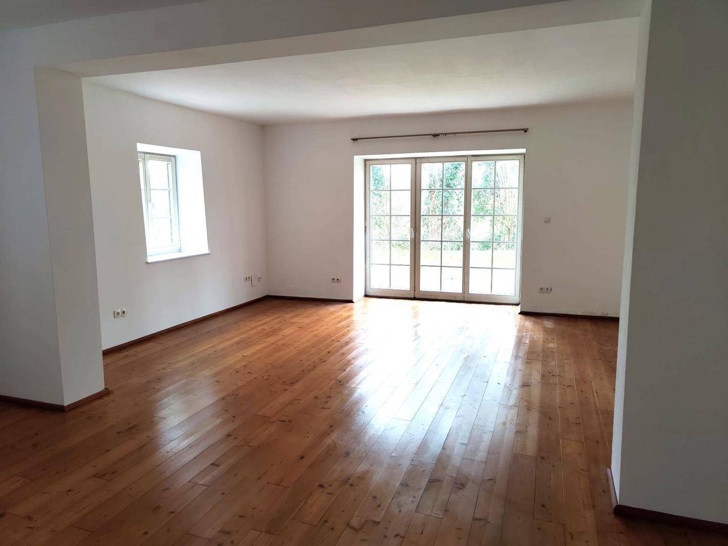 Helles, leeres Wohnzimmer mit Holzdielenboden und Zugang zum Garten durch große Türen.
