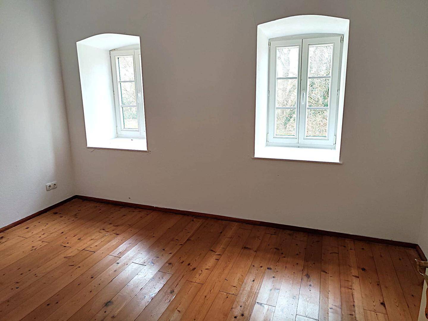 Helles Schlafzimmer mit Holzdielenboden und zwei Fenstern mit Blick ins Grüne.