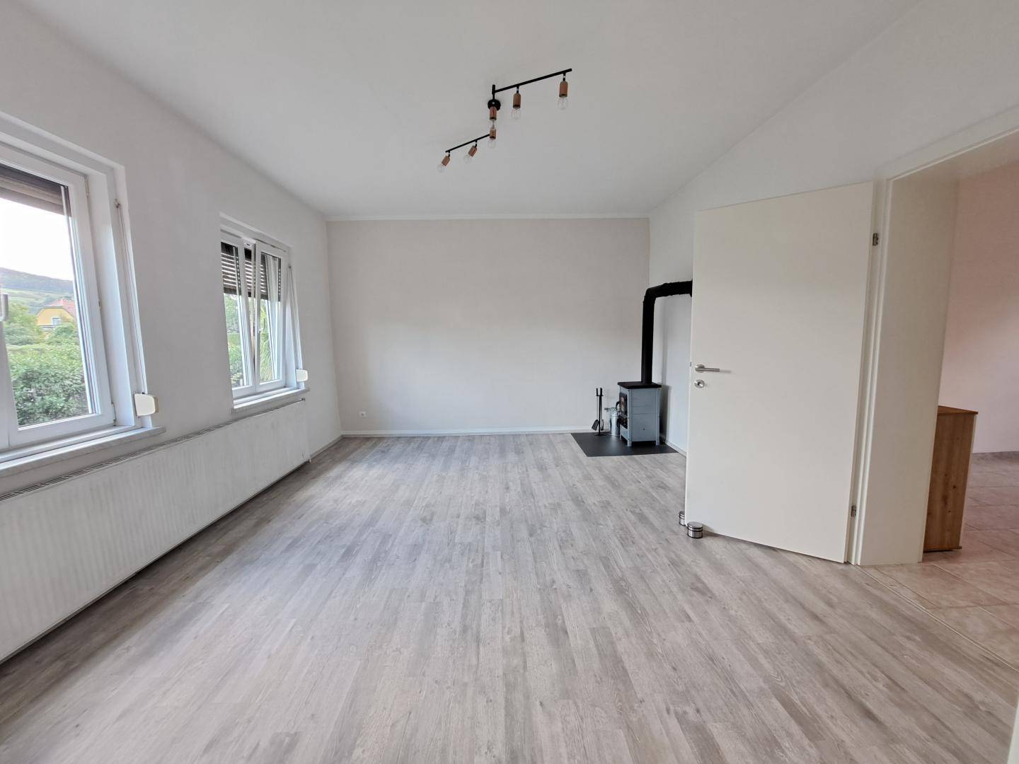 Wohnzimmer mit modernem Laminatboden, Kaminofen und zwei großen Fenstern.