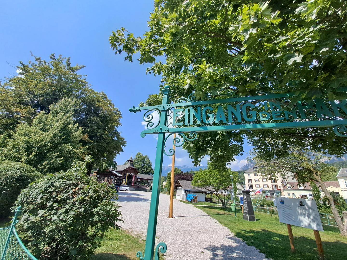 Eingangsbereich eines Parks oder Dorfplatzes mit Bäumen und Bergen im Hintergrund.