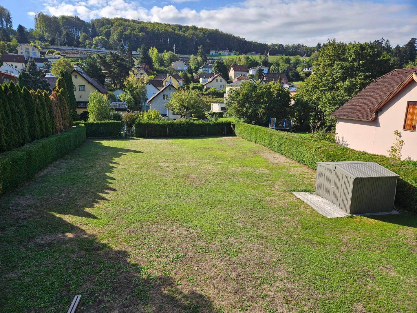 Weitläufiger Garten mit Rasenfläche, Geräteschuppen und Blick auf die umliegende Wohnsiedlung und Hügel.