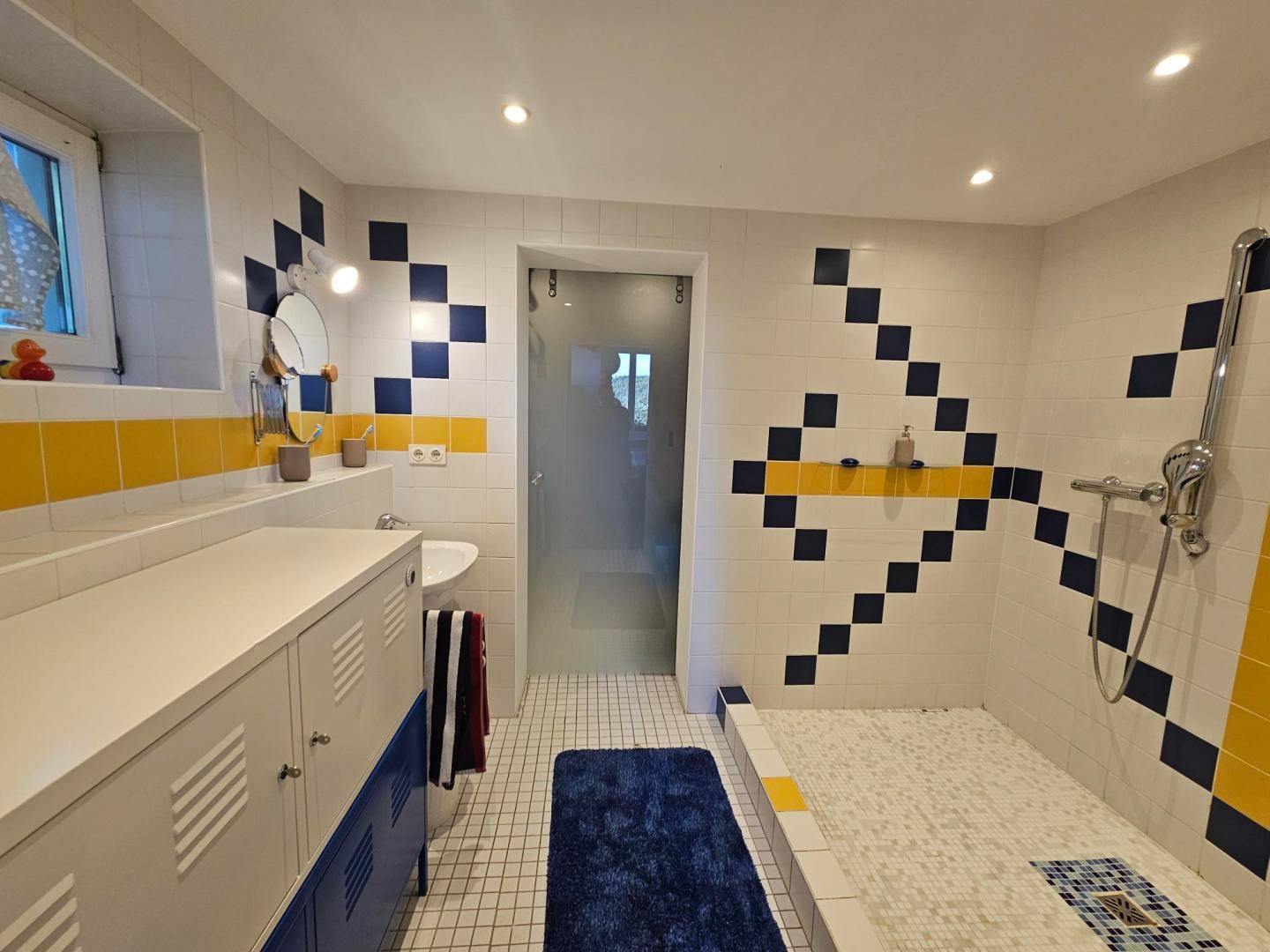 Badezimmer mit ebenerdiger Dusche, Waschbecken und Fliesen in Gelb, Blau und Weiß.