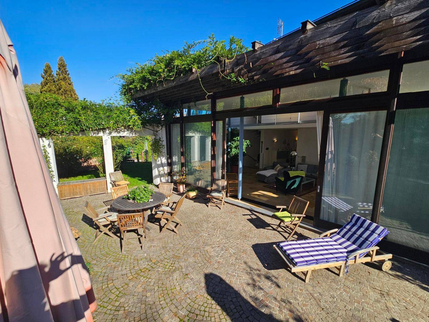 Sonniger Terrassenbereich mit Liegestuhl und Esstisch, angrenzend an die Glasfassade des Hauses.