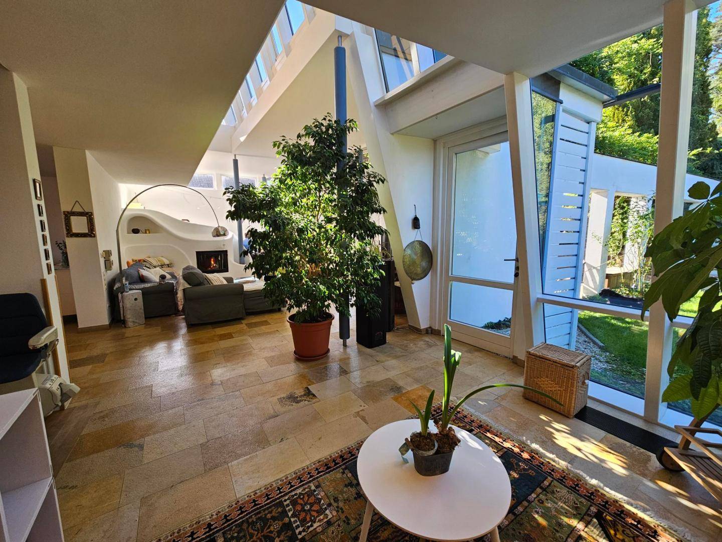 Heller Wohnbereich mit großen Fenstern, Steinboden und einem gemütlichen Kamin im Hintergrund.