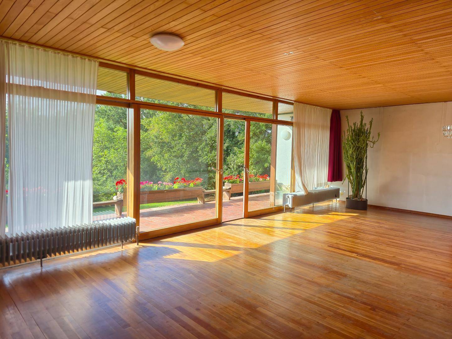 Helles Wohnzimmer mit Holzboden und Holzdecke, großen Fensterfronten und Blick ins Grüne.