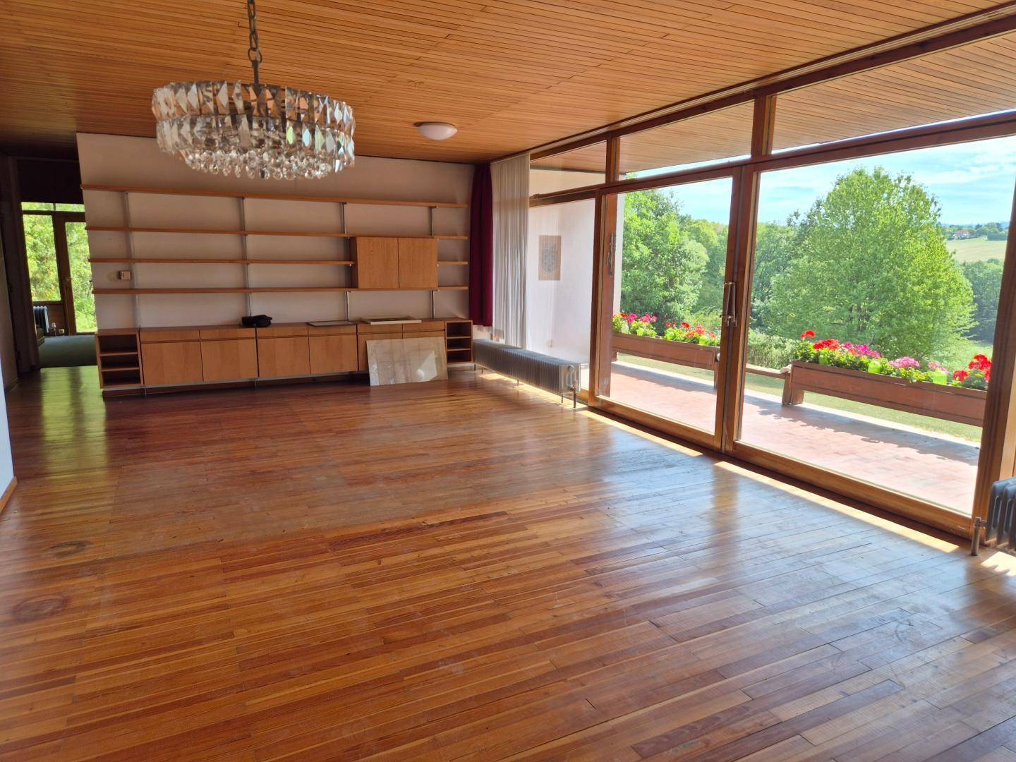 Geräumiges Wohnzimmer mit Holzboden, Holzdecke und eingebauten Regalen, bietet einen weiten Blick ins Grüne.