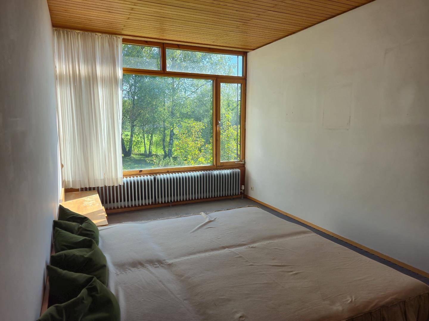 Schlafzimmer mit Holzboden, Holzdecke und großen Fenstern, die viel Tageslicht und einen Blick ins Grüne bieten.