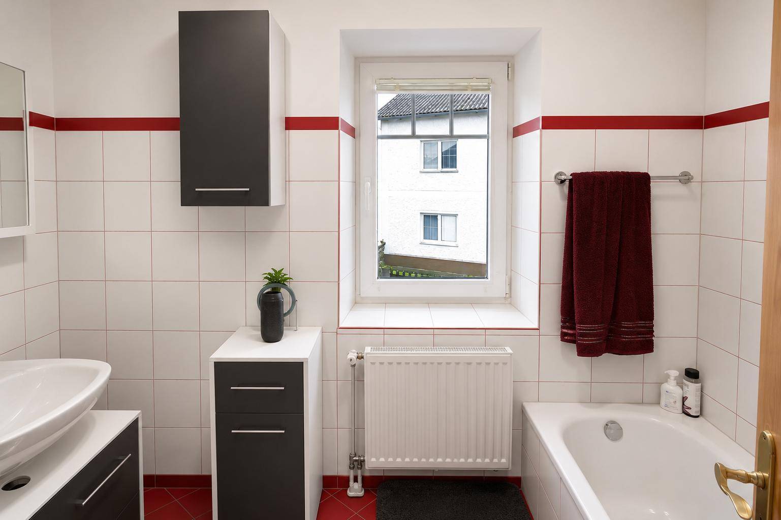Helles Badezimmer mit Badewanne, Waschbecken, Fenster und roten Akzentstreifen.