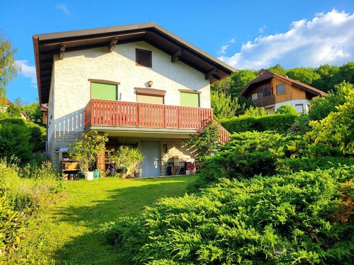 Hausfassade mit Balkon und grünen Fensterläden, umgeben von einem gepflegten Garten und Sträuchern.