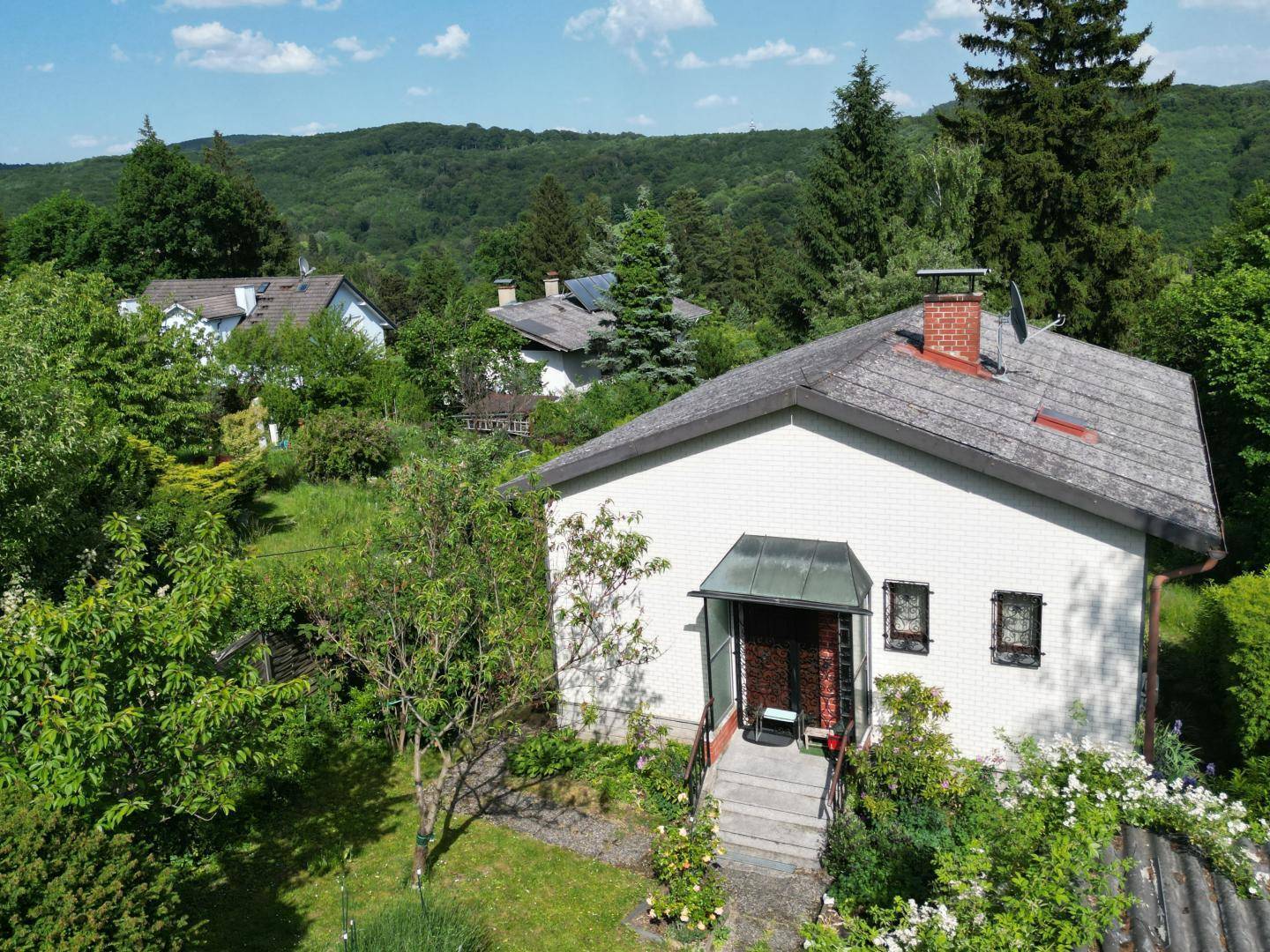 Luftaufnahme eines Hauses mit Satteldach und grüner Umgebung, umgeben von Bäumen und Hügeln.