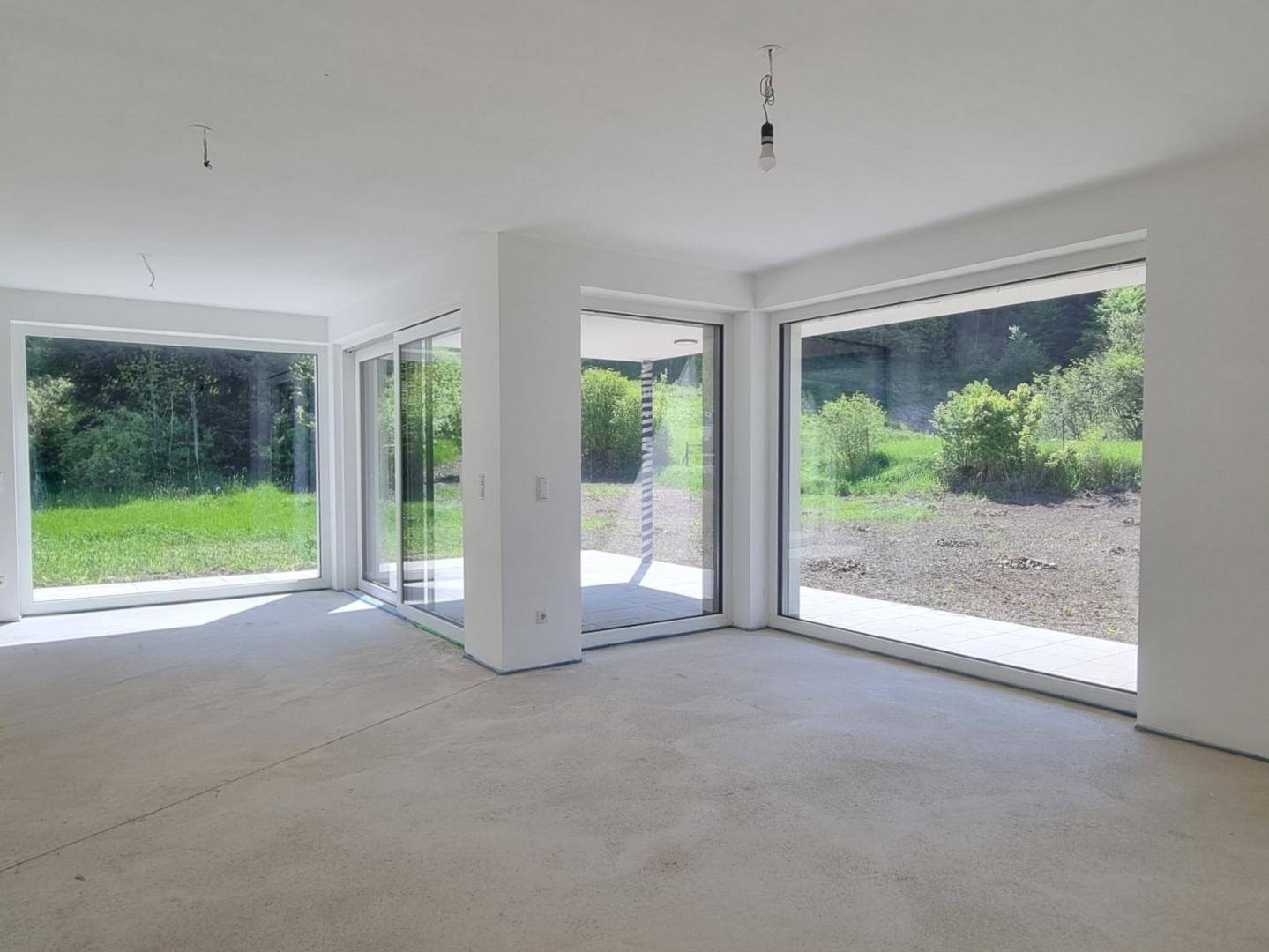Offener Wohnbereich im Rohbau mit mehreren großen Fenstern und Zugang zur Terrasse.