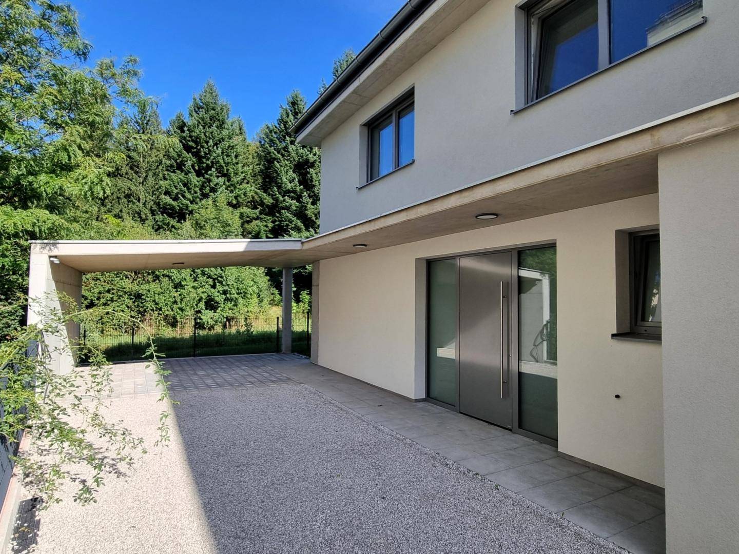 Moderne Hausfassade mit großem Glaseingang, Kiesauffahrt und einem überdachten Carport im Grünen.