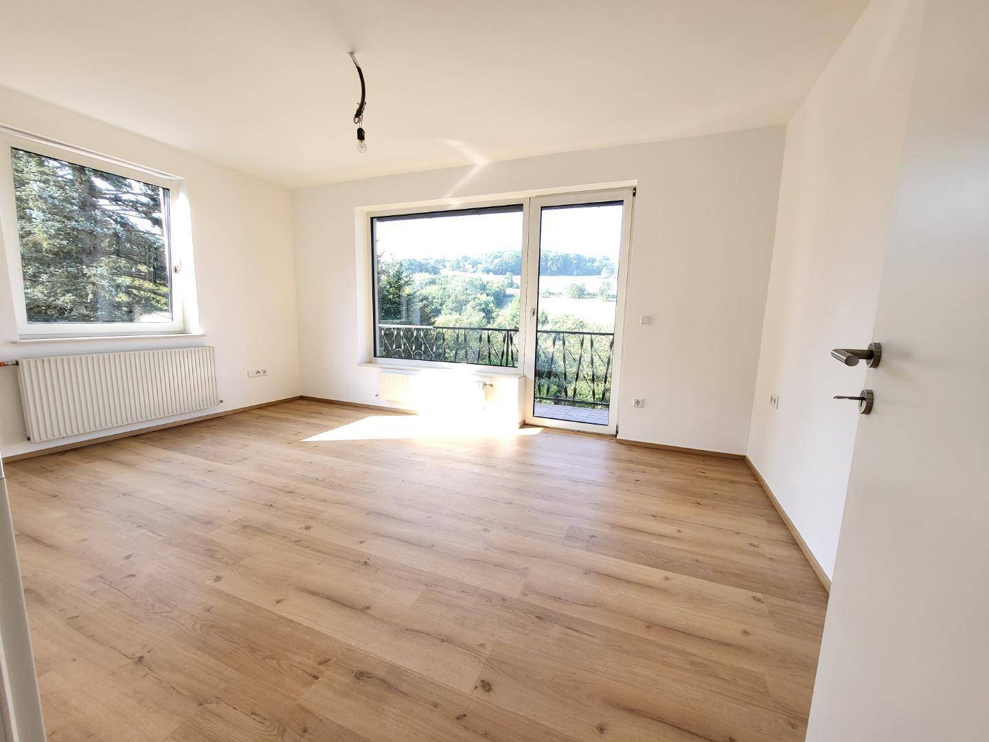 Helles, leeres Zimmer mit Laminatboden, großen Fenstern und Zugang zum Balkon.