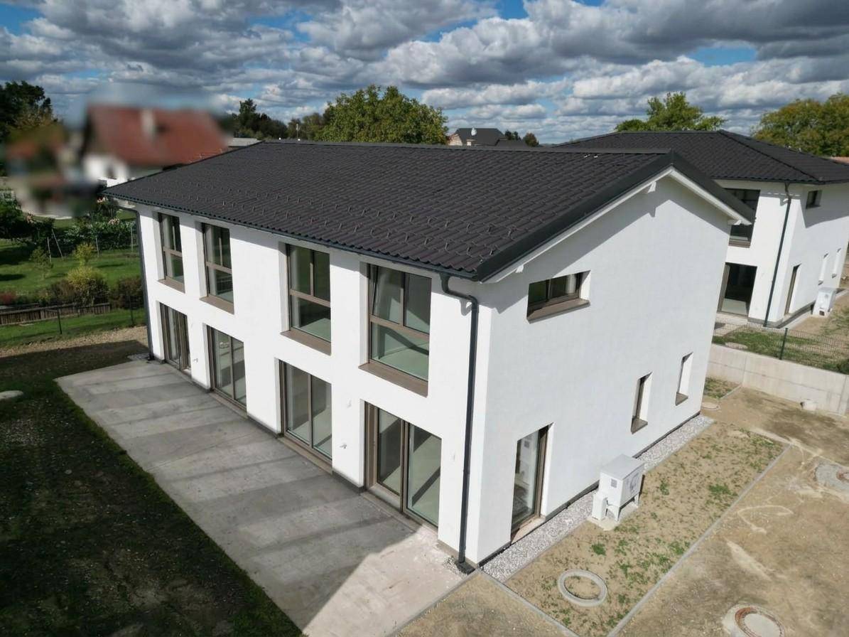 Neubau-Doppelhaushälfte mit weißer Fassade und dunklem Ziegeldach in ländlicher Umgebung.