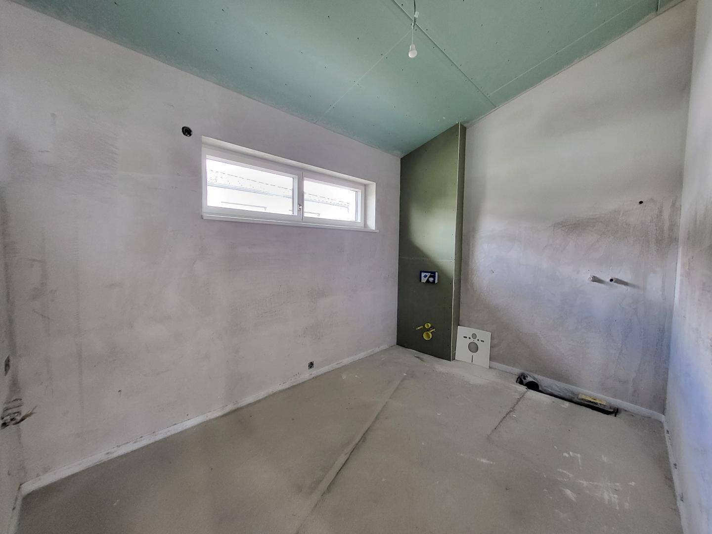 Badezimmer im Rohbau mit Fenster und vorbereiteten Anschlüssen für Dusche, Waschbecken und Toilette.