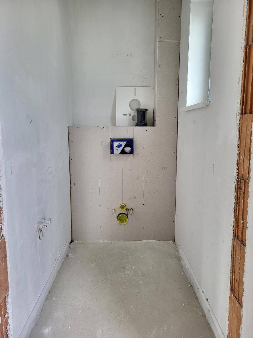 Unfertiges Gäste-WC mit vorbereiteten Anschlüssen für Toilette und Waschbecken sowie einem kleinen Fenster.