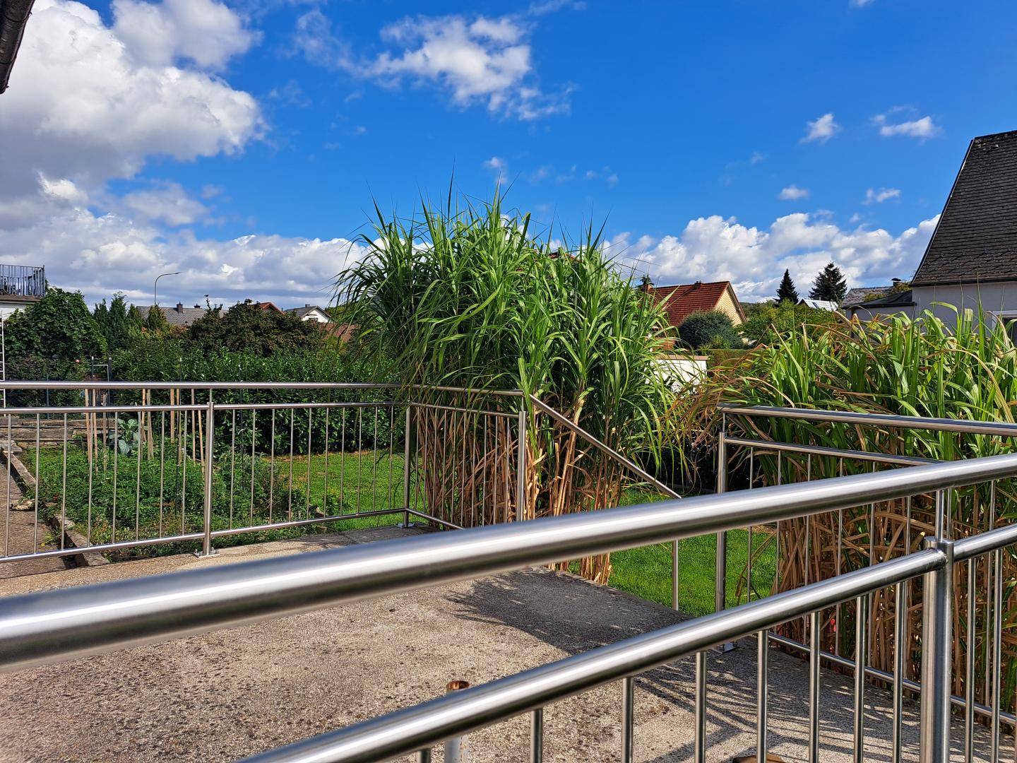 Terrasse mit Metallgeländer und Blick ins Grüne unter blauem Himmel.