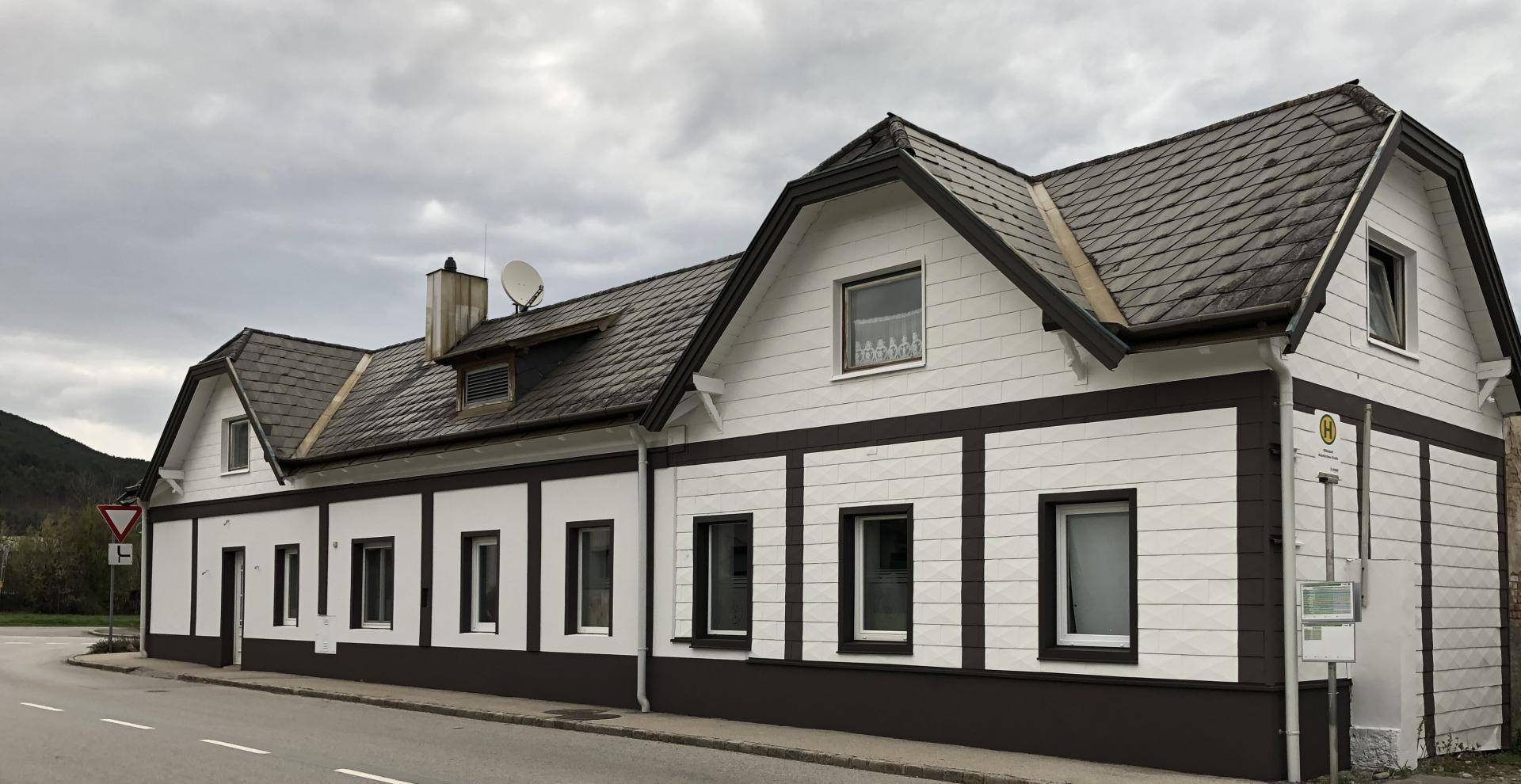 Traditionelles Gebäude mit weißer Holzfassade und dunklem Dach, gelegen an einer ländlichen Straße.