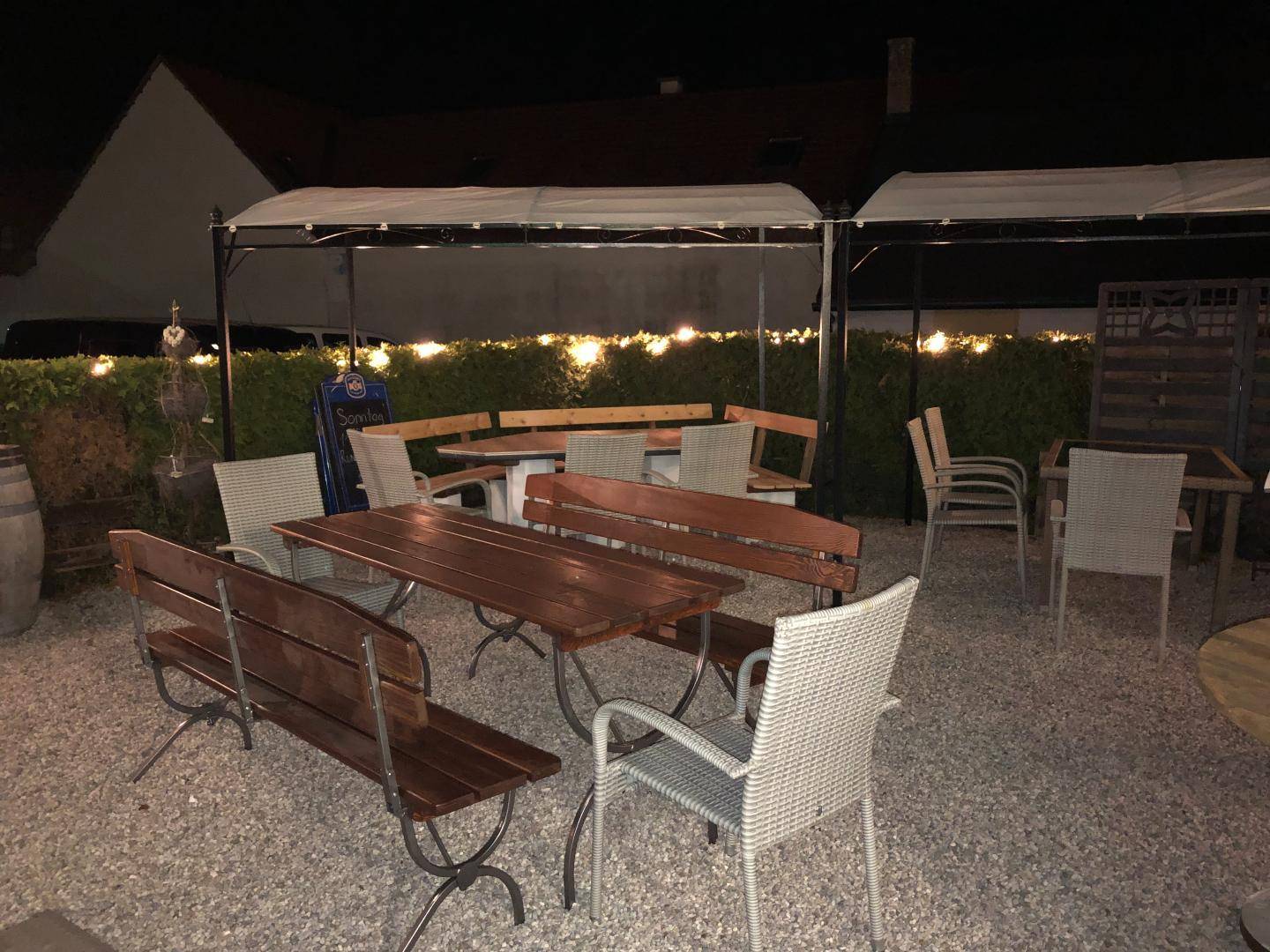 Beleuchteter Außenbereich am Abend mit mehreren Sitzgruppen und Pergolen, umgeben von Hecken.