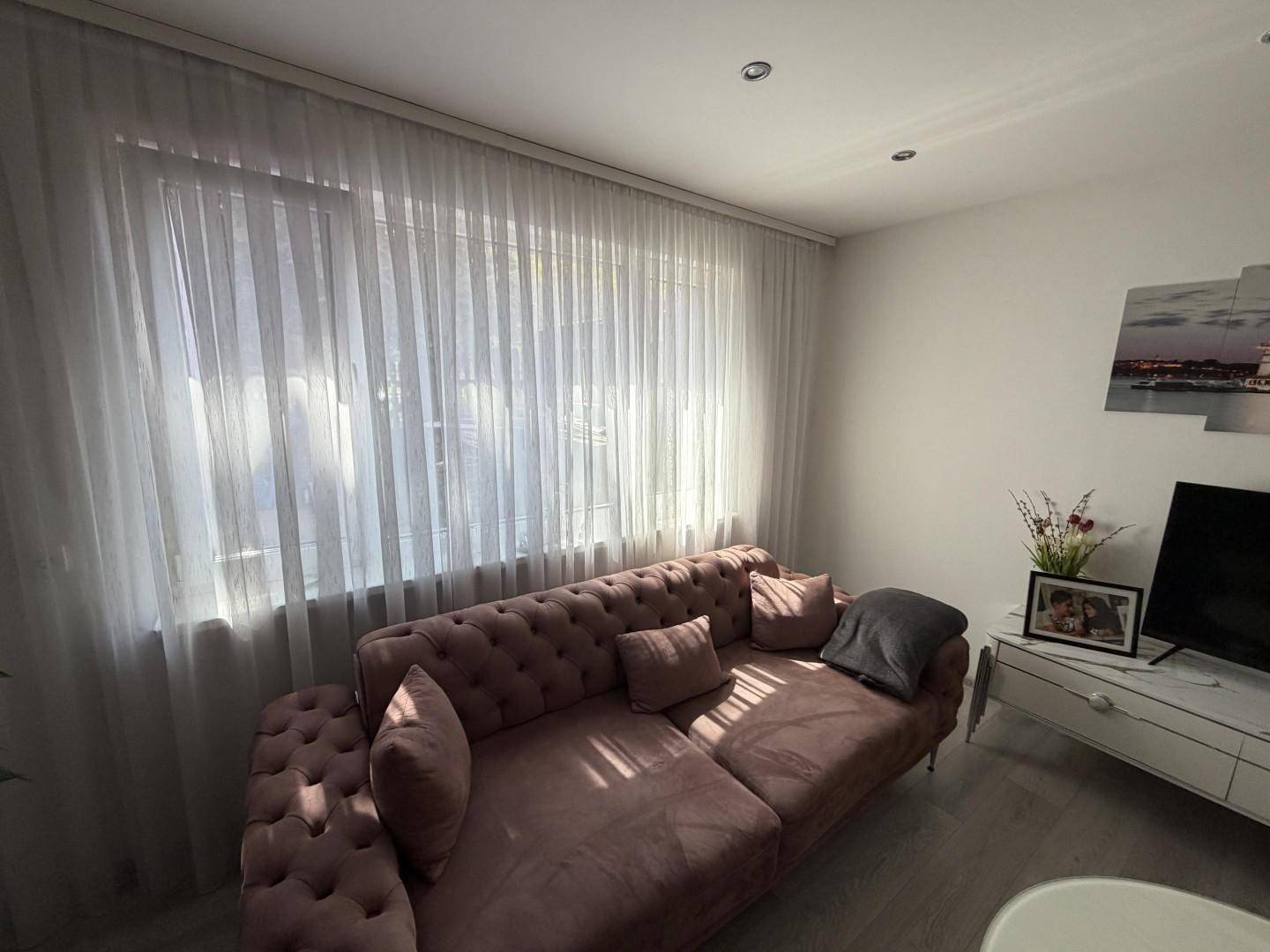 Lichtdurchflutetes Wohnzimmer mit Vorhängen, bequemer Couch und hellem Boden.