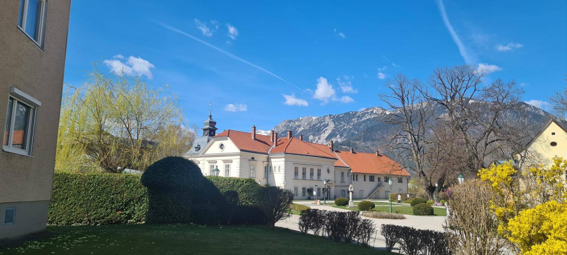 Historisches Gebäude mit rotem Dach und Bergkulisse, umgeben von grünen Hecken und Bäumen.