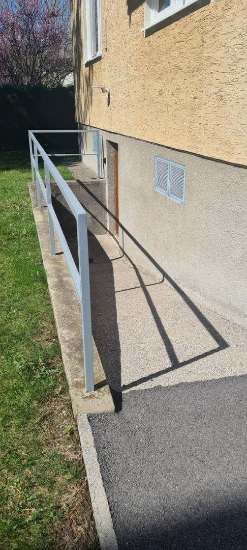 Barrierefreie Rampe mit Handlauf führt zu einem Eingang an der Gebäudeseite.