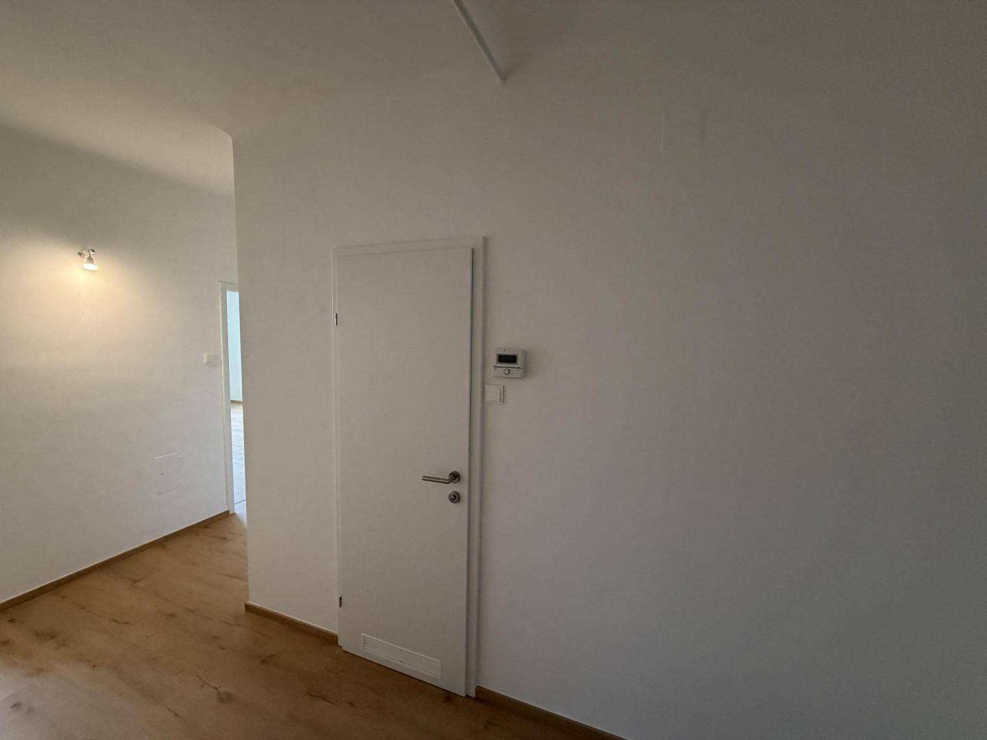 Heller Flur mit Holzboden, weißer Tür und einem Thermostat an der Wand.