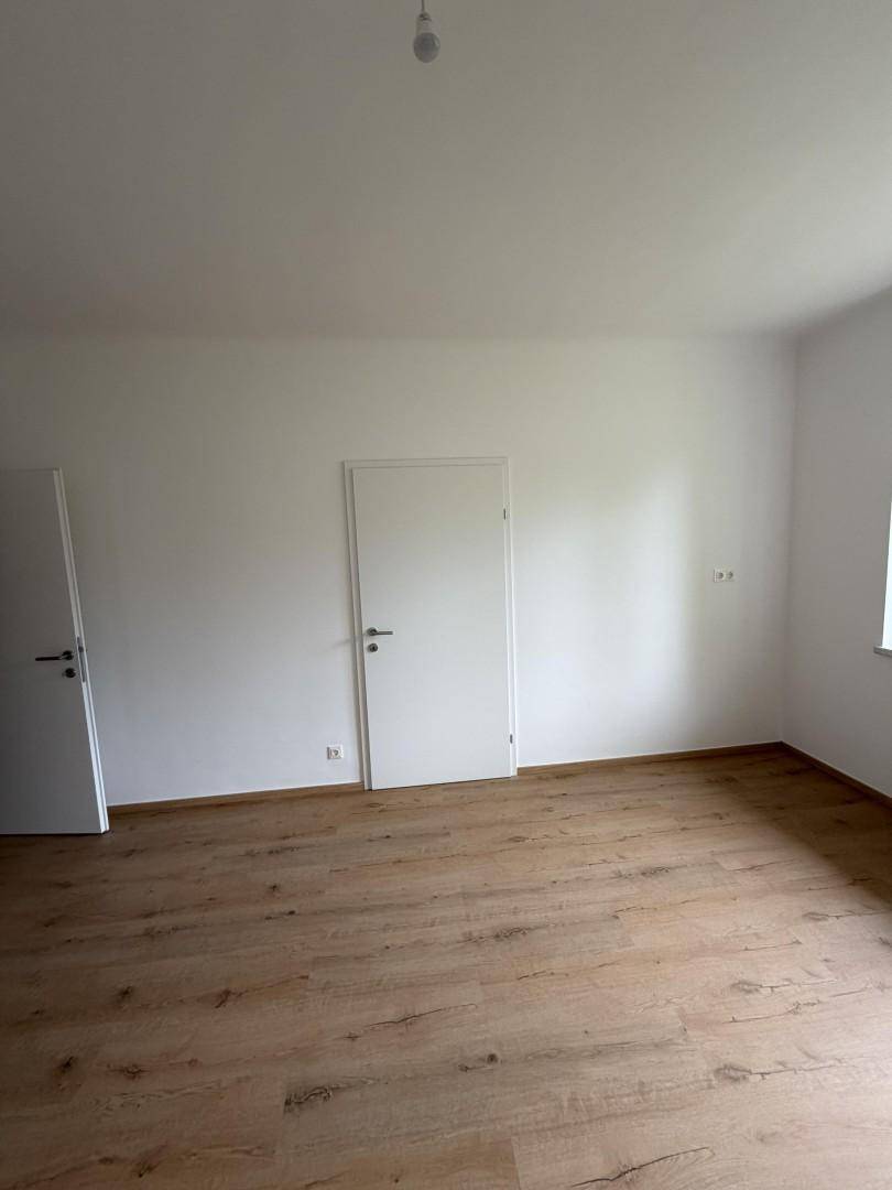 Helles, leeres Zimmer mit Holzboden und zwei weißen Türen, bereit zur individuellen Gestaltung.