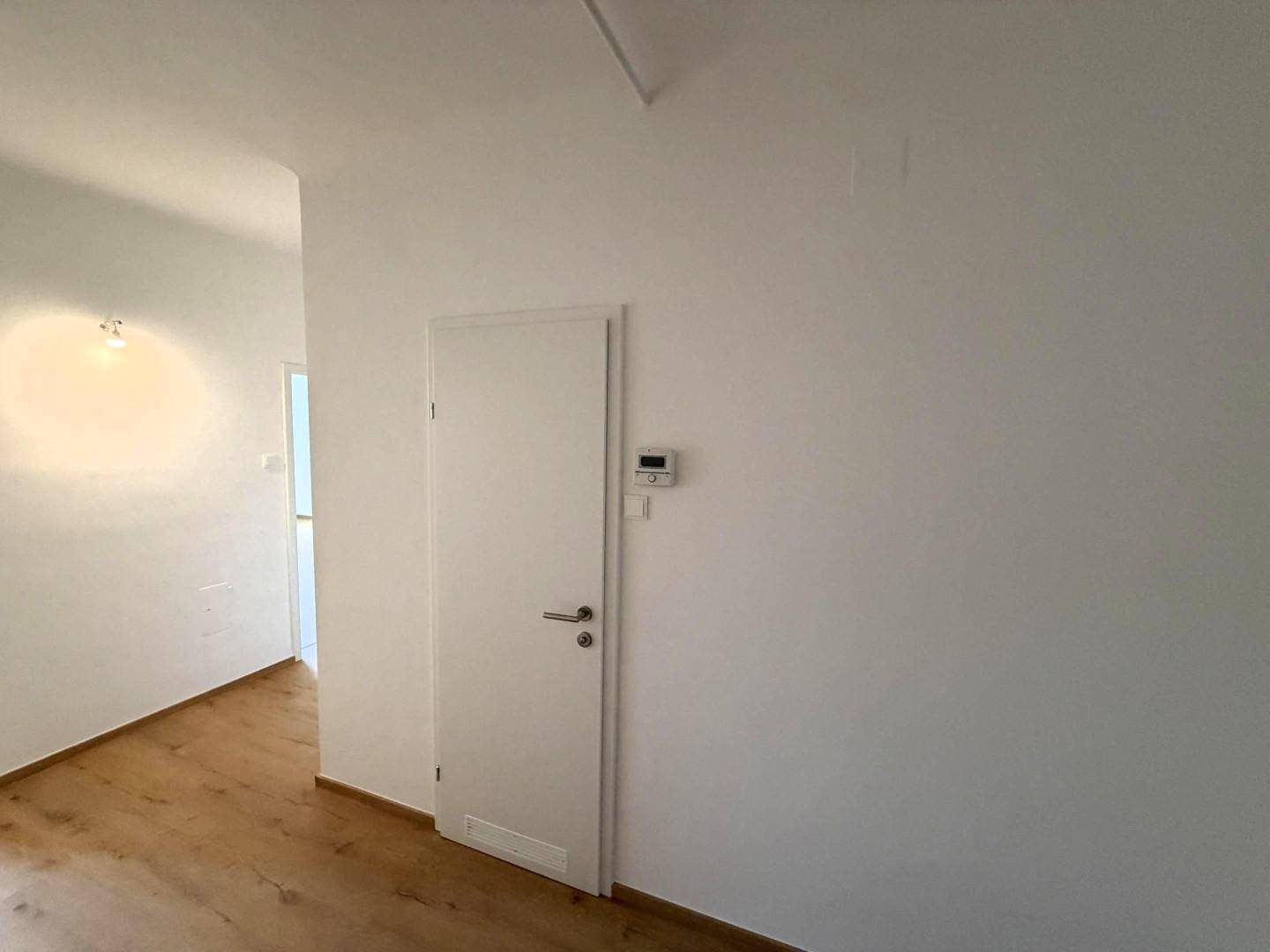 Heller Flur mit weißer Tür, Holzboden und einem Heizungsthermostat an der Wand.
