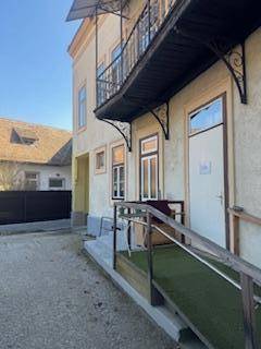 Eingangsbereich eines Gebäudes mit einer Rollstuhlrampe und einem Balkon im Obergeschoss.