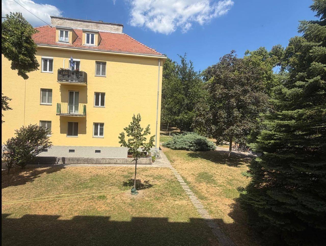 Gelbes Mehrfamilienhaus mit rotem Ziegeldach und Balkonen, umgeben von einer grünen Gartenanlage.
