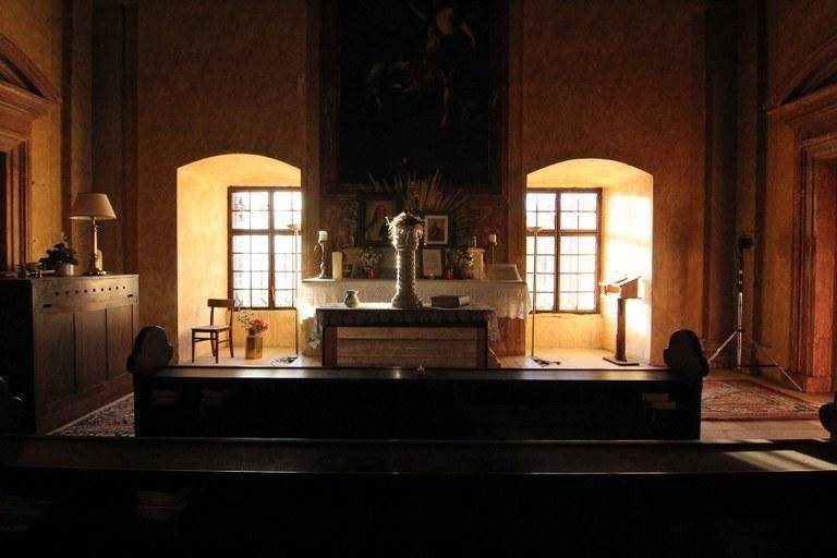 Dämmeriger Innenraum einer Kapelle mit Kirchenbänken, einem Altar und zwei hell erleuchteten Fenstern.